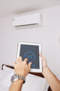 A pair of hands holding an electronic tablet device using a smart thermostat app to adjust the temperature of a mini-split air conditioner on the wall of a bedroom.