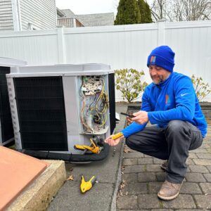 repair man crouching down next to an exterior air conditioning unit that is being repaired.