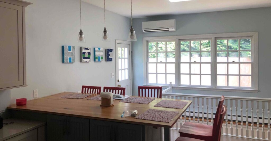 A mini-split air conditioning unit mounted a the wall above a window in a home's kitchen and dining area.