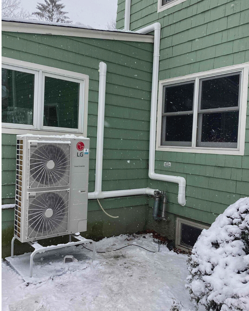 Ductless heating unit installed near the ground outside of a green-colored residential home with snow on the ground.