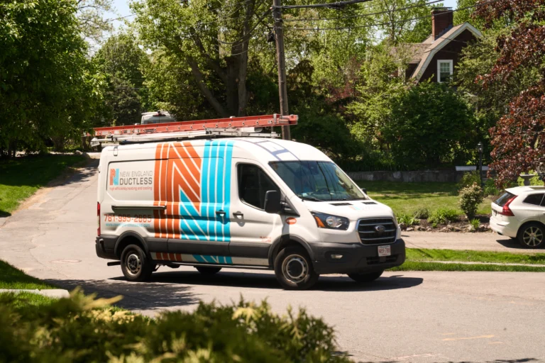 A New England Ductless service van, specializing in A/C repair, is parked on a suburban street beside trees and houses, with a red ladder mounted on the roof.