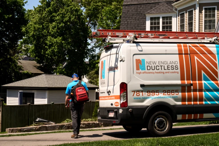A technician carrying a red backpack walks toward a house next to a New England Ductless service van parked on a suburban street.