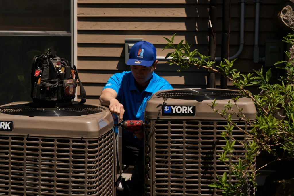 A technician in a blue shirt and cap performs A/C Repair on one of two York air conditioning units outside a building, using a power tool. A tool bag rests on top of one unit.