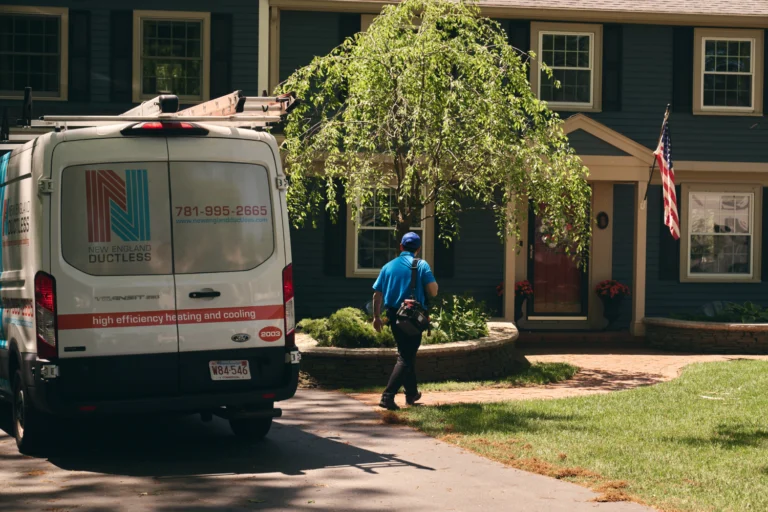 A technician specializing in A/C repair walks toward a blue house from a New England Ductless service van parked in the driveway. An American flag is mounted near the entrance.