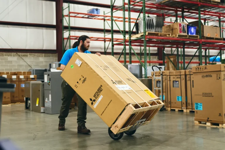 A man in a warehouse moves a large boxed item on a hand truck. Industrial shelving and other packages are visible in the background.