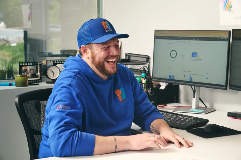 A man in a blue hoodie and cap sits at a desk smiling, with two computer monitors displaying graphs and data charts in the background.