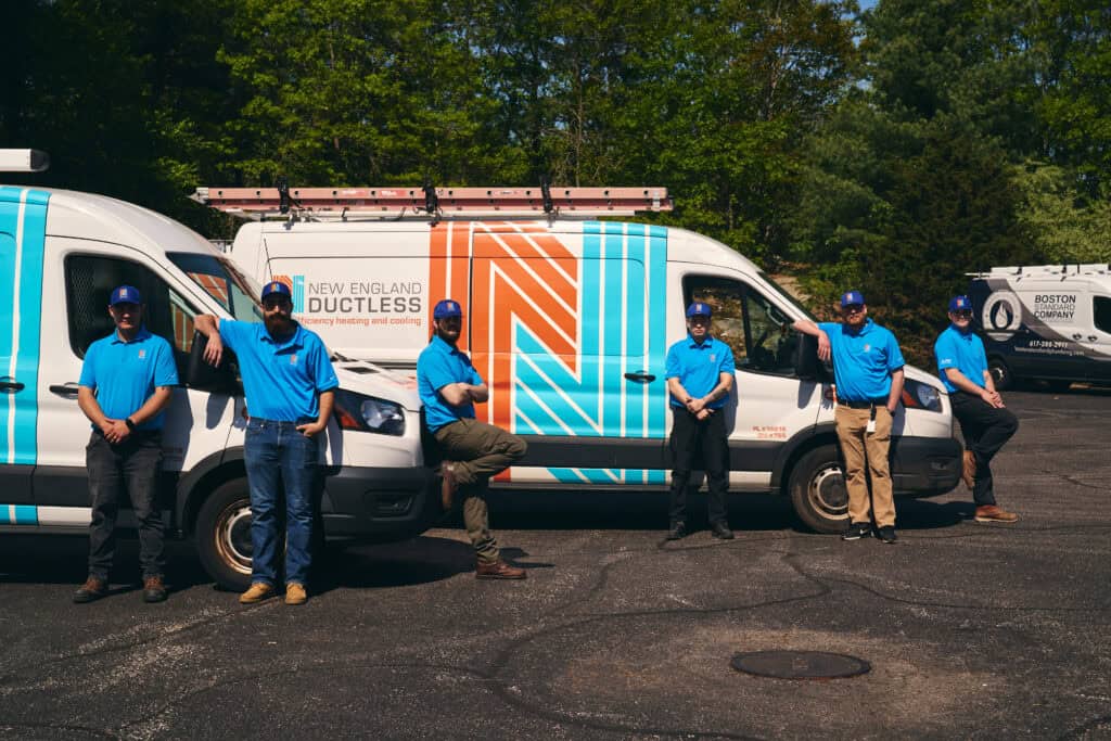 Six workers in blue uniforms stand beside two branded vans labeled “New England Ductless” in a parking lot, with trees and another company van in the background.