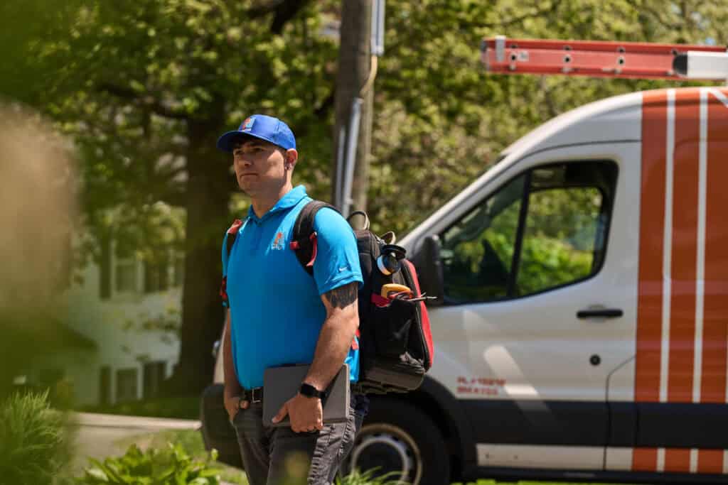 A technician wearing a blue uniform and cap stands outdoors carrying tools and a tablet, with a white utility van parked nearby.