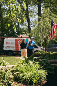 A technician talks with a woman in a yard near a New England Ductless service van; an American flag is visible in the background.