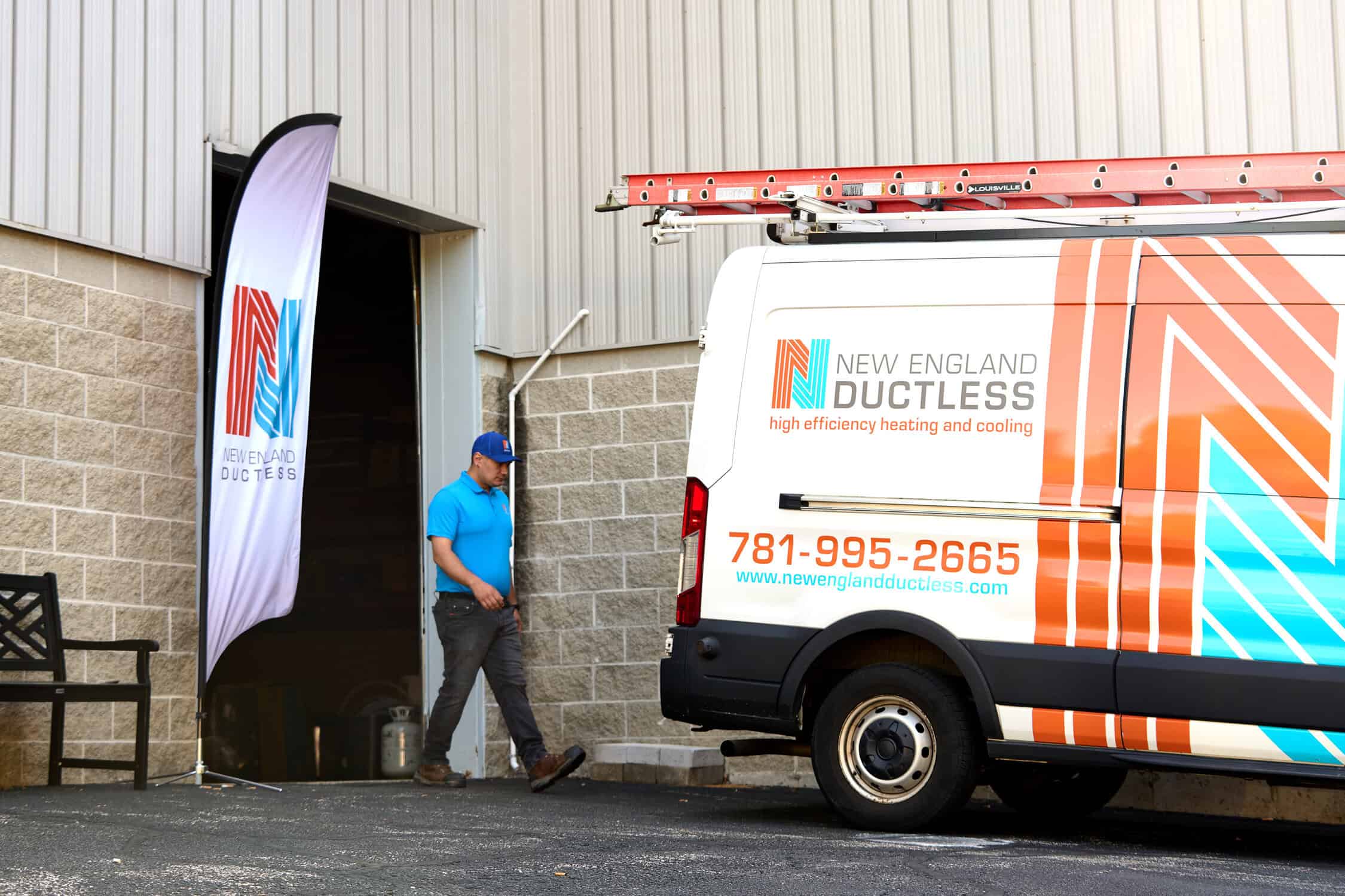 A technician walks into a building next to a New England Ductless service van branded with its company logo and phone number.