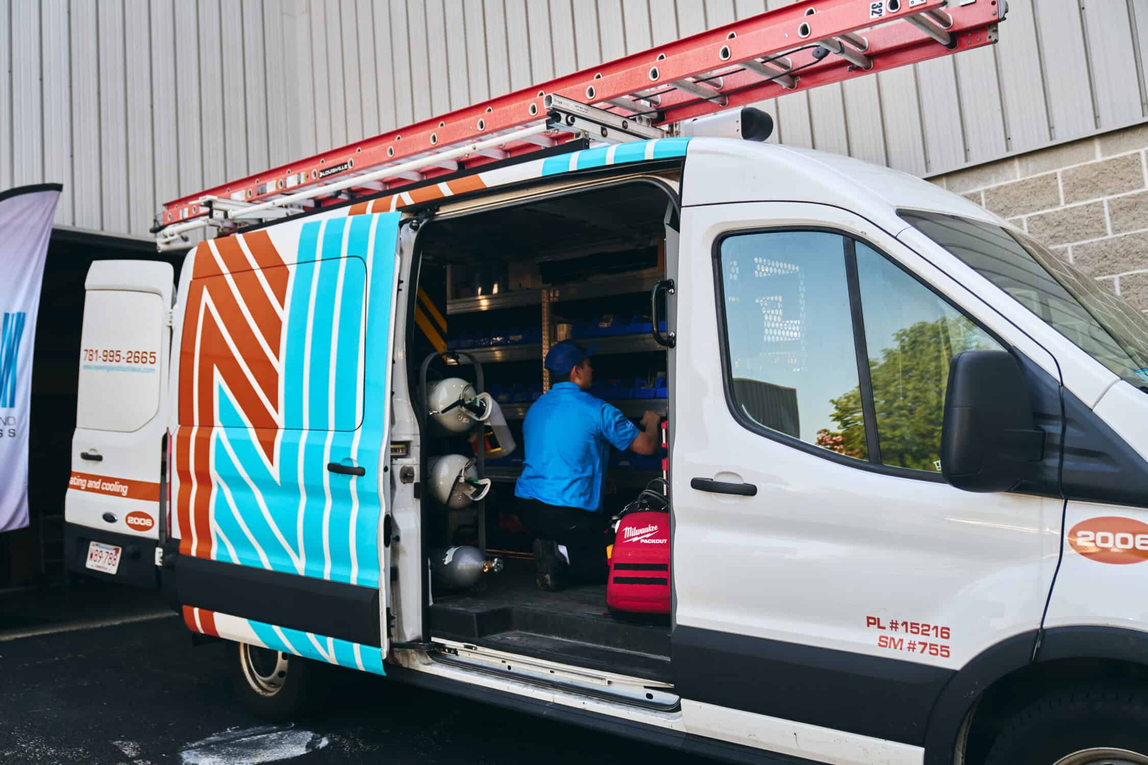 A technician in a blue shirt works inside an open utility van equipped with tools and shelving, parked beside a building.