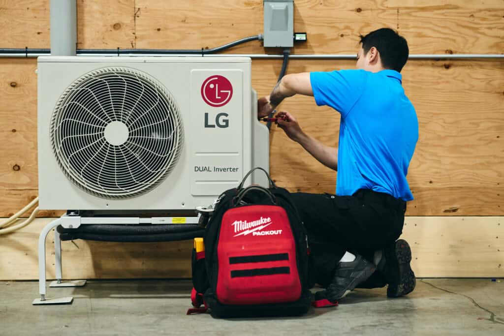 A technician in a blue shirt repairs an LG outdoor HVAC unit, with a Milwaukee tool bag sitting on the floor beside them.