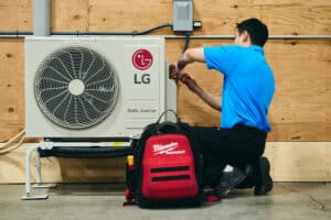 A technician in a blue shirt repairs an LG outdoor HVAC unit, with a Milwaukee tool bag sitting on the floor beside them.