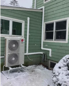 Outdoor LG air conditioning unit installed against a green-sided house in winter, with snow on the ground and falling. White piping runs along the exterior wall, showcasing reliable heating services even in cold conditions.